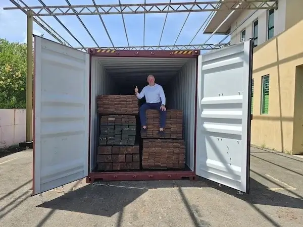 Man sitting on stacked timber inside an open shipping container, giving thumbs up